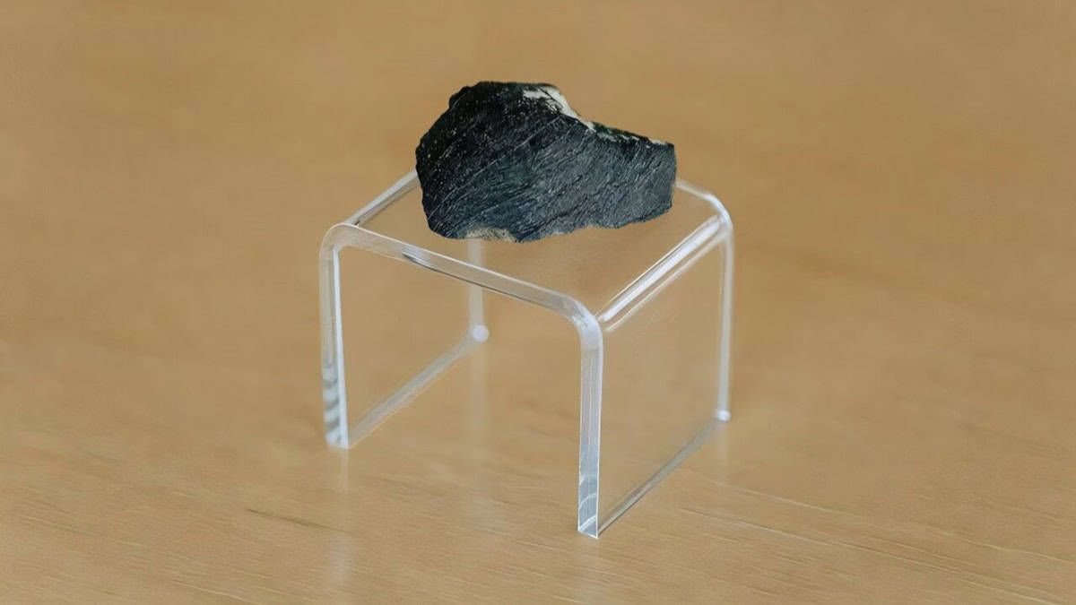 A small black rock meteorite on see-through plastic stand against wooden background