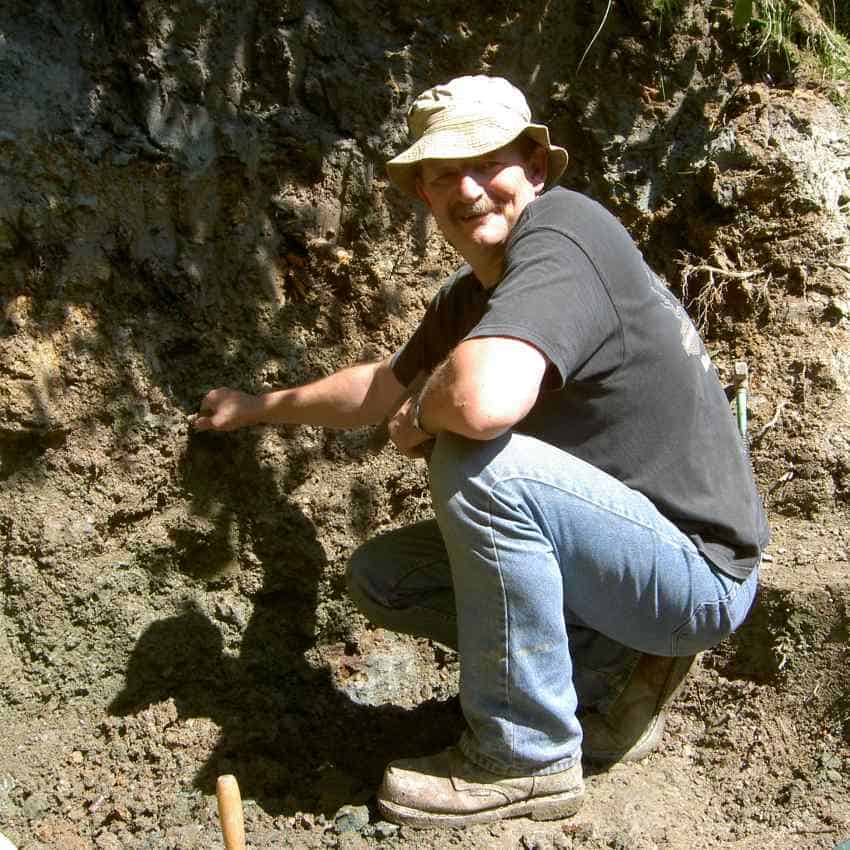 A smiling man squats by a rock face.