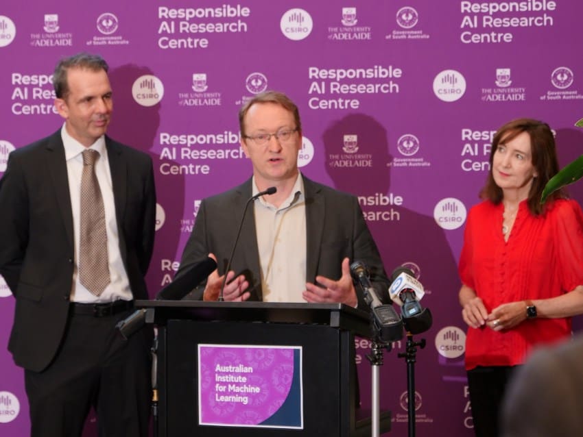 3 individuals stand behind a podium. There are 2 men wearing dark grey suits and a woman wearing a bright red blouse and black pants.