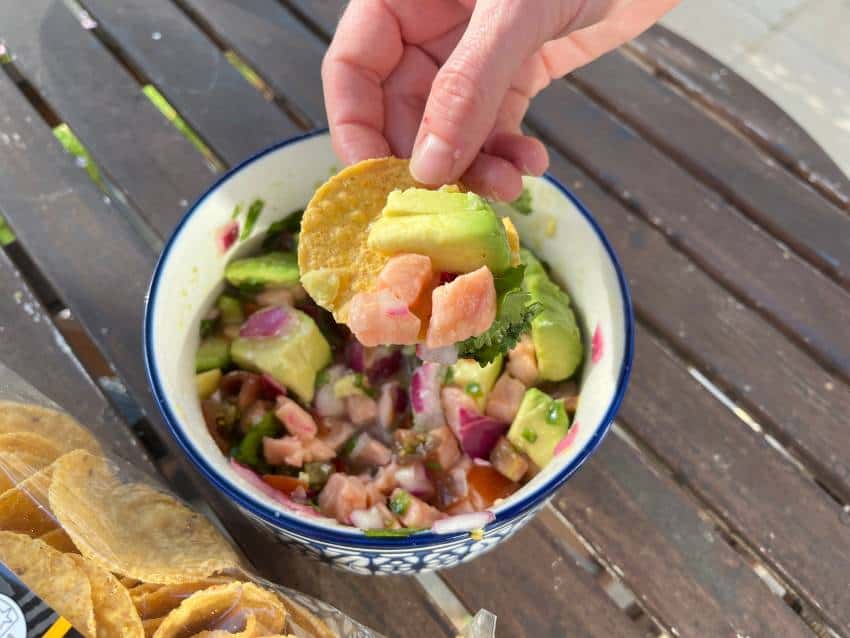 A hand holding a corn chip scoops pink fish and avocado from a bowl containing ceviche.