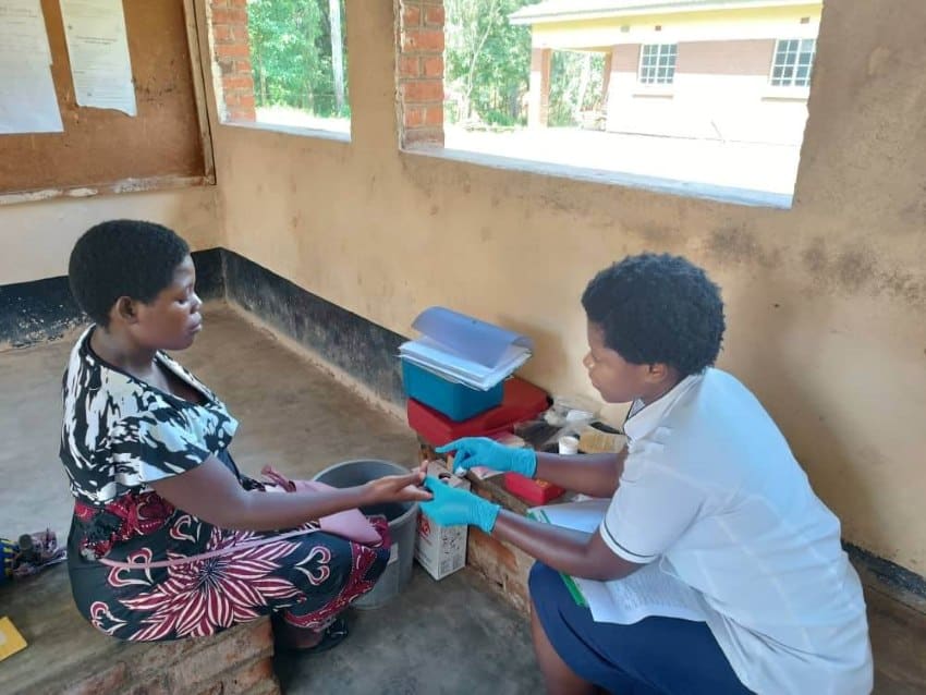 Two women sit on the floor of a building. One is wearing gloves and is taking a sample of blood from the other, who is pregnant.