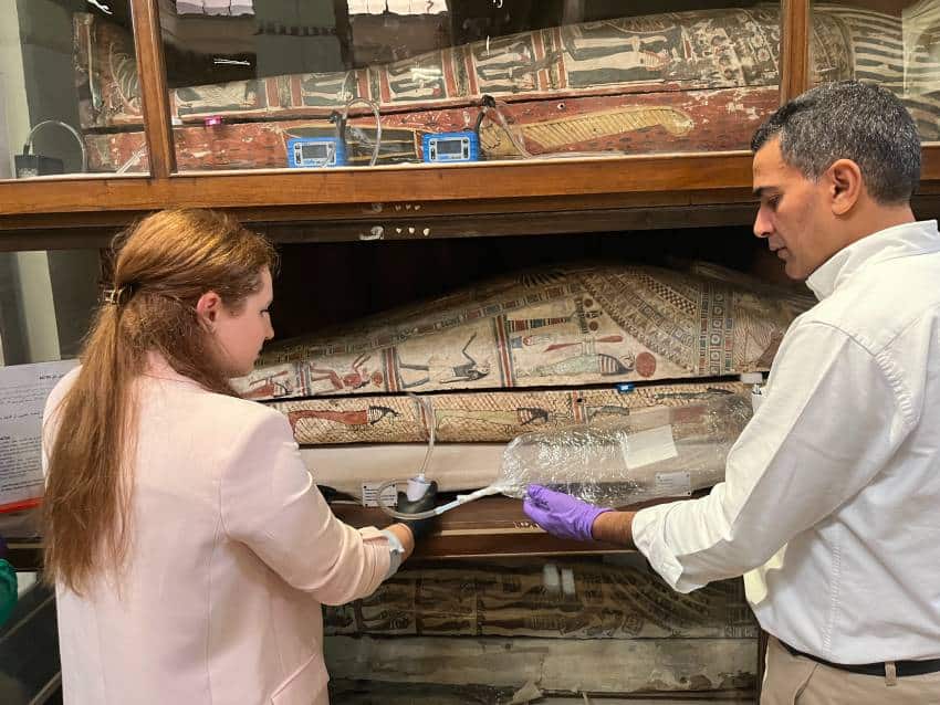 Two scientists, one female and one male use equipment near an ancient egyptian mummy to discern its smell.