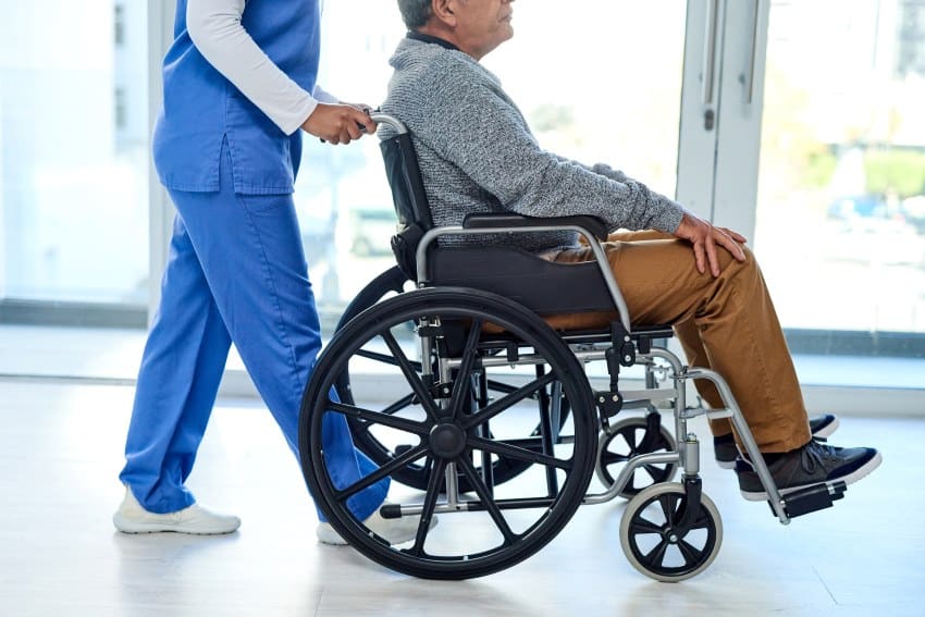 A older male patient sitting in a wheelchair is being pushed by a health professional in blue scrubs