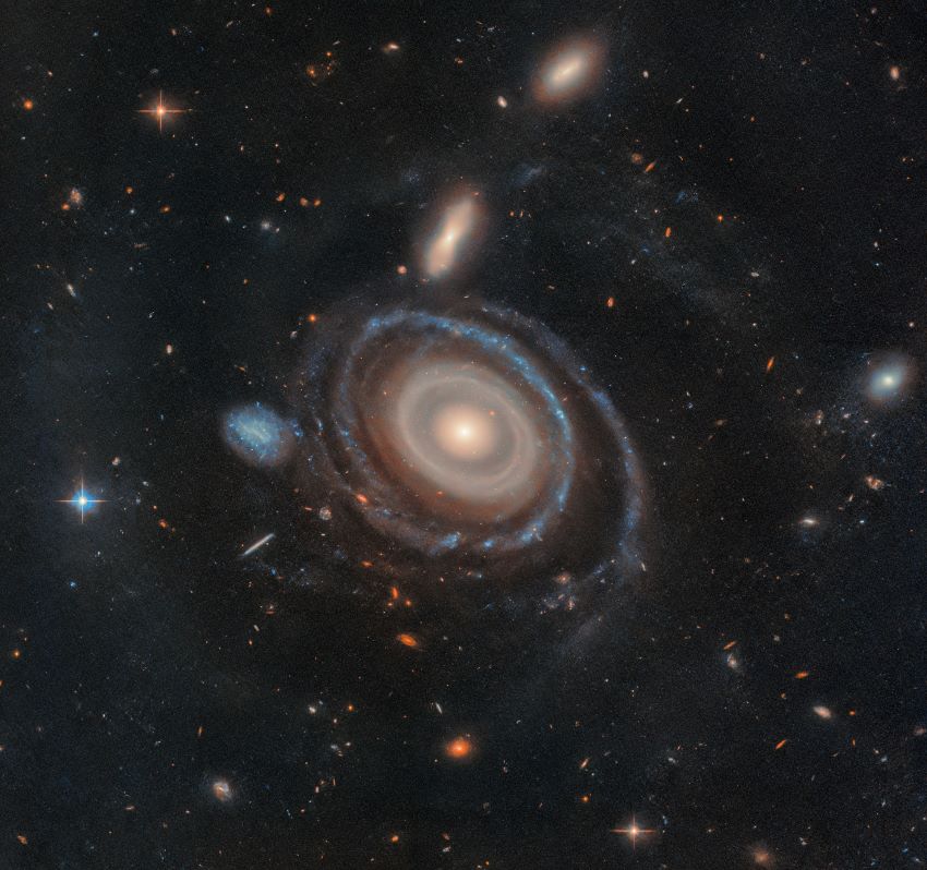 A photograph of a galaxy surrounded by several glowing rings. Next to it is a small blue galaxy. They are surrounded by many stars and distant galaxies
