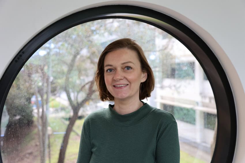 A woman with short brown hair, wearing a green long-sleeved top is standing in front of a circular window