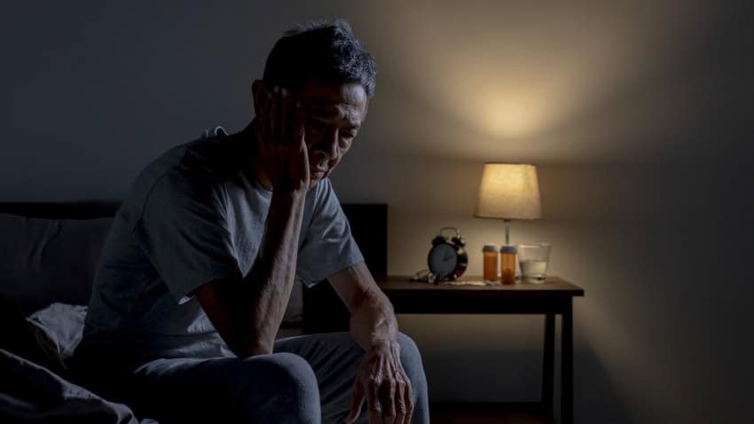 An older asian mad sits on the edge of his bed with his head in his hand. In the background there are pill bottles on his bedside table.