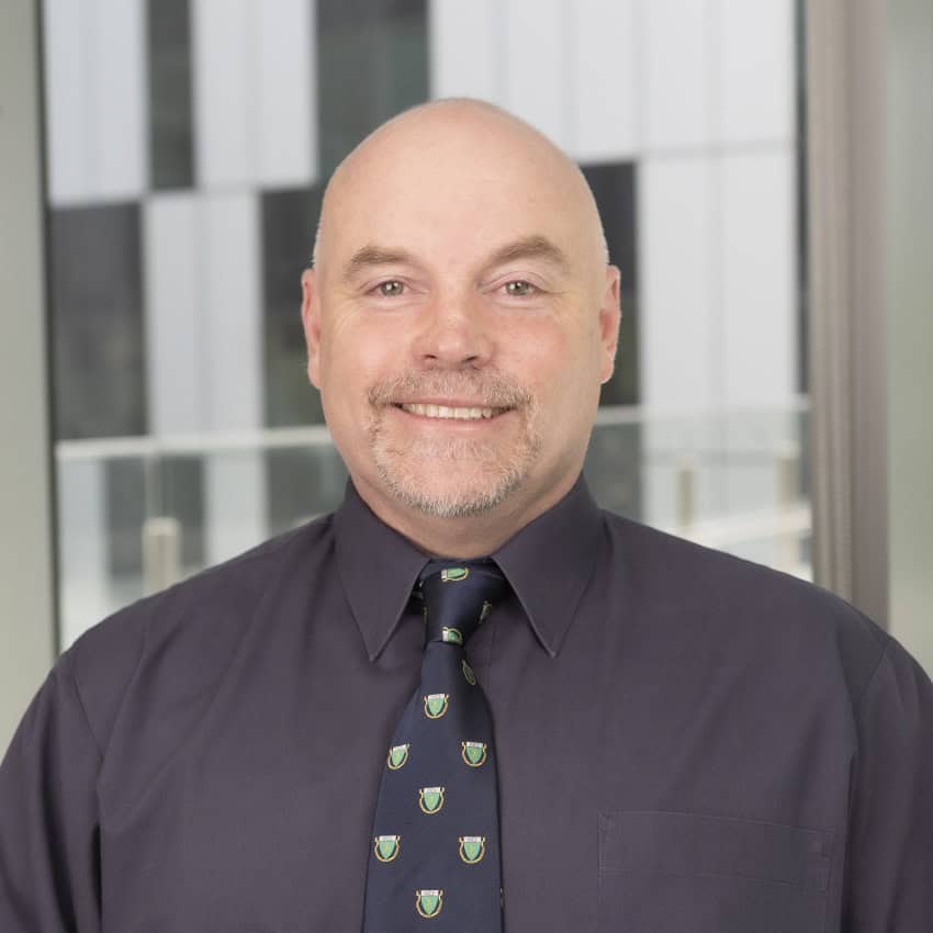 A man with a beard and bald head smiles at the camera. He is wearing a dark purple shirt and tie