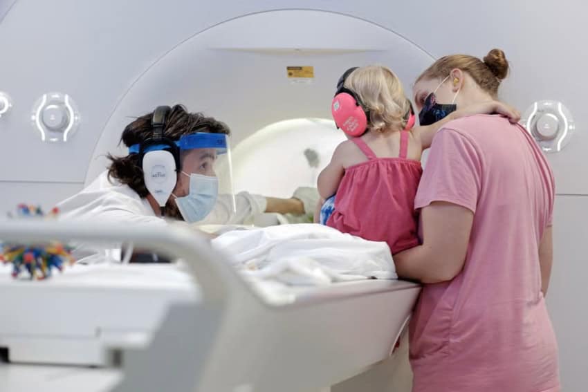 A man wearing a ppe crouches next to a young girl wearing pink earphones, who is being held by a woman wearing a pink t-shirt. They are in front of a mri machine