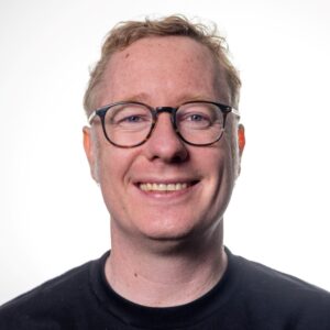 A man with blonde hair and round glasses smiles at the camera against a white background