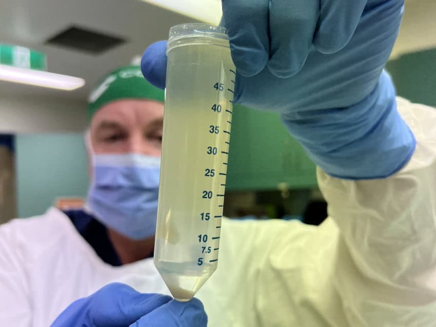 A man wearing surgical gown, mask, hair cover, and gloves holds a test tube full of a murky liquid