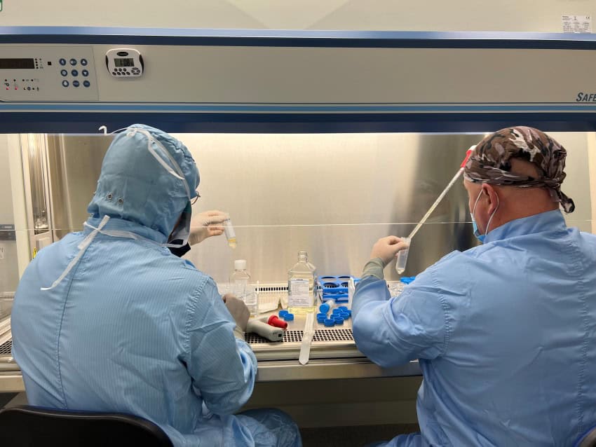2 people dressed in blue ppe sit at a fume hood while they do biological experiments