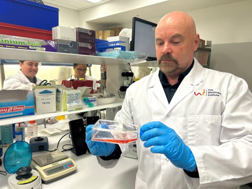 A man wearing a white lab coat and blue gloves stands in a laboratory with 2 team members seen on the other side of some lab benches. He is holding a clear container with pink liquid inside