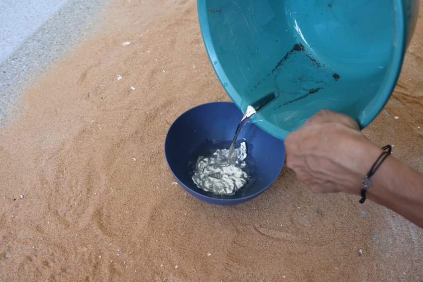 A someone off camera pours liquid mercury and water from a large blue bucket into a smaller one, leaving behind dirt and debris