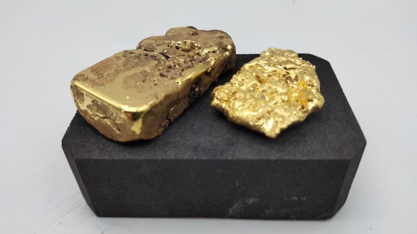 Two irregular nuggets of gold sit atop a black block against a white background