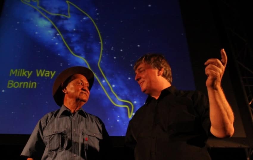 A photo of 2 men standing in front of a projection of the milky way with the outline of a bird superimposed in yellow. On the left is wardaman elder yidumduma bill harney wearing a short sleeve shirt and hat. On the right is astrophysicist roy norris wearing a black shirt
