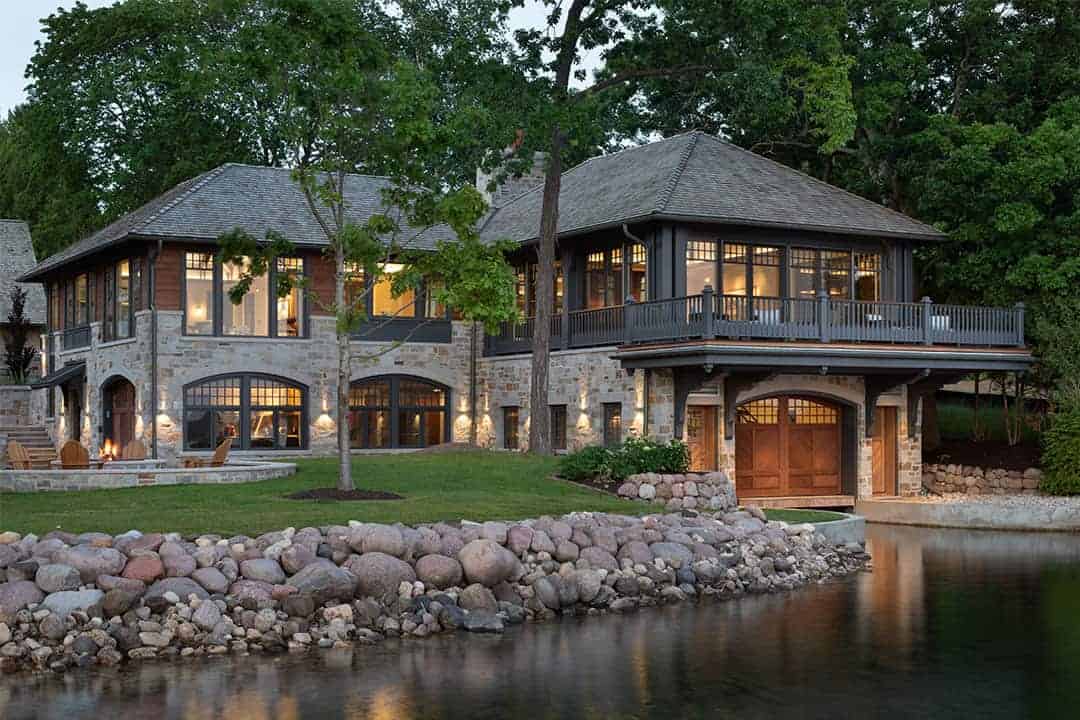 Twilight view of a stone lakefront boathouse with an arched wooden boathouse door beneath a covered upper-level deck, surrounded by lush landscaping and an outdoor fire pit lounge; warm interior lighting glows through arched windows, set along a boulder-lined shoreline with mature trees framing the property.