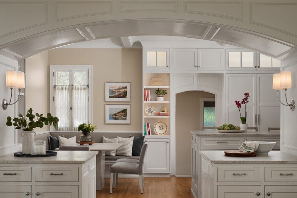 Classic white kitchen viewed through a grand coffered archway, featuring twin marble-topped islands, glass-front upper cabinetry, and built-in bookshelves; a cozy breakfast nook with upholstered banquette seating and a rustic wood table sits beneath a warm gallery of framed landscape prints.
