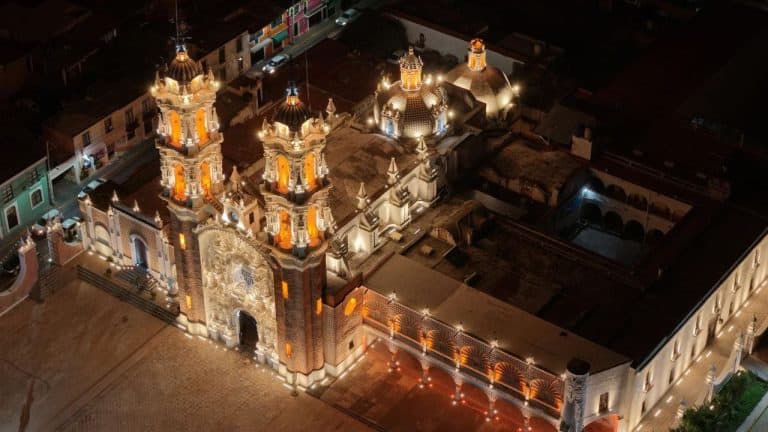 Imágen aérea de una iglesia iluminada por la noche en un entorno urbano en Tlaxcala, México. La iglesia presenta arquitectura colonial con torres y cúpulas decoradas con iluminación cálida y destacada, resaltando en el cielo nocturno.