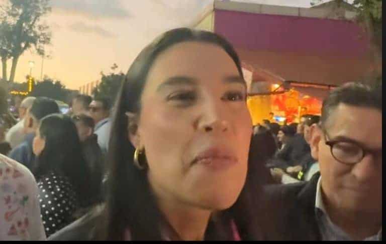 María Acosta en un evento público, rodeada de muchas personas, en un entorno al aire libre durante el atardecer, mostrando una expresión seria y segura, con fondo ilustrando actividad social.