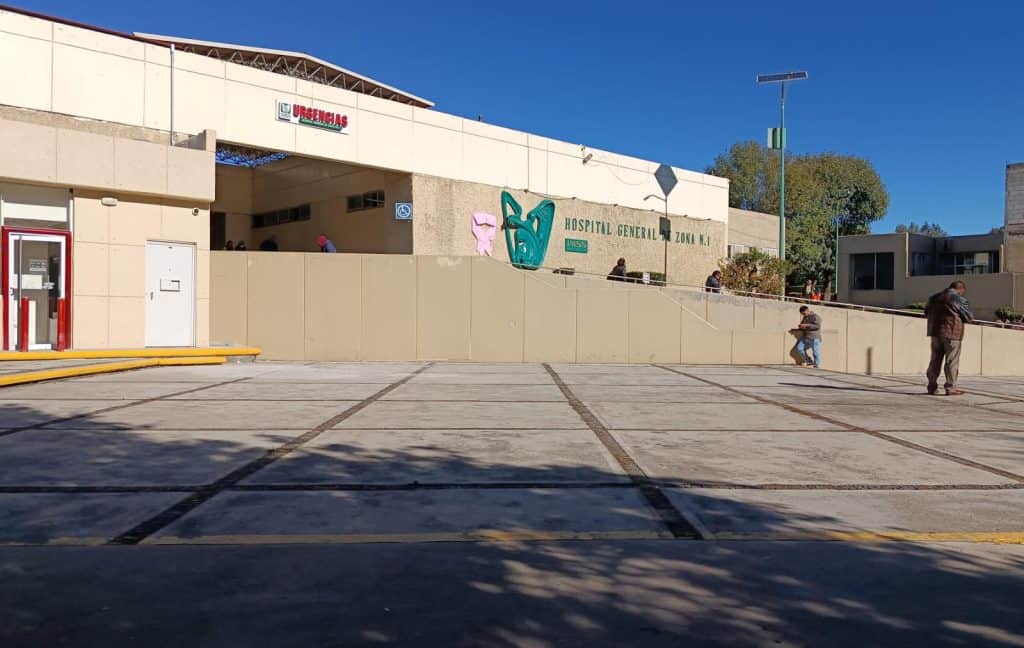 Hospital general de Tlaxcala en un día soleado, con personas esperando afuera y algunas utilizando sus dispositivos móviles. El edificio cuenta con un acceso principal visible y un cartel que indica que es un hospital público.
