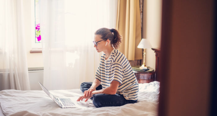 Young woman with glasses sitting on the bed at home using a laptop to communicate online work and education