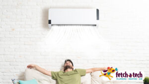 A man enjoys relaxing on his couch as cool, refreshing air flows from a wall-mounted air conditioner. The Fetch‑A‑Tech logo in the corner shows this is a family-owned Las Vegas company dedicated to keeping your home comfortable with honest, helpful service.