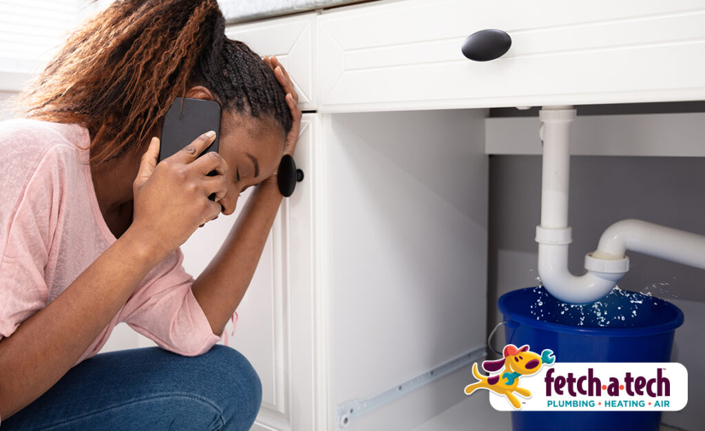 A concerned woman in Las Vegas speaks on the phone while kneeling beside a leaking pipe under her sink, catching dripping water in a blue bucket. The Fetch‑A‑Tech Plumbing logo is displayed, highlighting our family-owned team’s commitment to providing helpful, dependable service and peace of mind when you need it most.