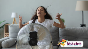 A woman in a white t-shirt sits in front of a large fan, looking overheated. The Fetch‑A‑Tech Plumbing, Heating, and Air logo appears in the bottom right corner, highlighting our commitment to keeping Las Vegas families cool and comfortable with honest, family-owned service you can trust.