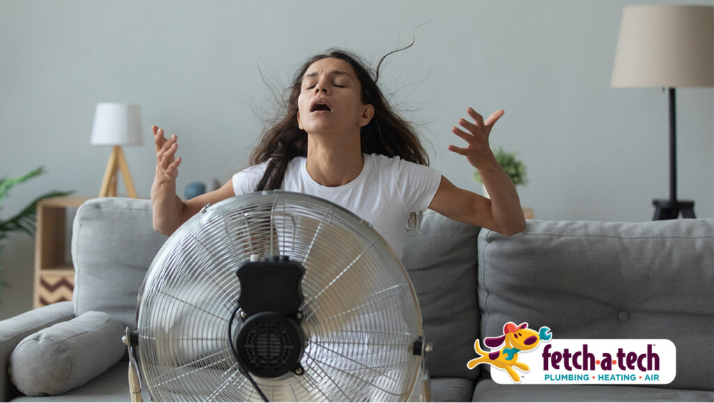 A woman in a white t-shirt sits in front of a large fan, looking overheated. The Fetch‑A‑Tech Plumbing, Heating, and Air logo appears in the bottom right corner, highlighting our commitment to keeping Las Vegas families cool and comfortable with honest, family-owned service you can trust.