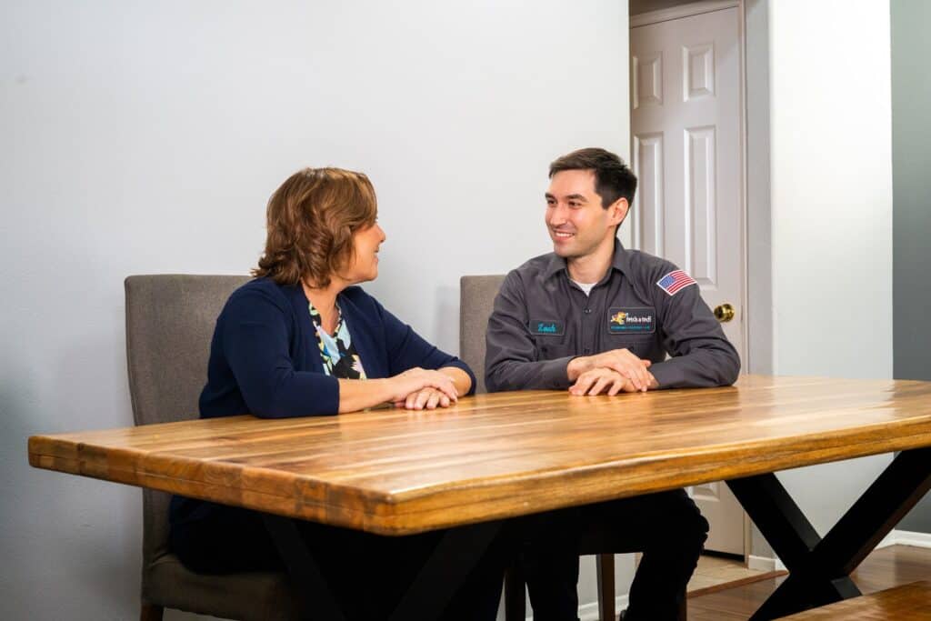 A friendly Fetch‑A‑Tech technician sits with a homeowner at a cozy wooden table, thoughtfully explaining insulation options to help keep her Las Vegas home comfortable and energy efficient. The relaxed home setting highlights our honest, family-owned approach and commitment to providing helpful service every step of the way.
