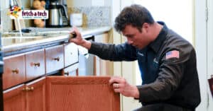 A friendly Fetch‑A‑Tech plumber in uniform carefully inspects the pipes beneath a kitchen sink, ensuring everything is safe and in good working order for a Las Vegas home. The Fetch‑A‑Tech logo is visible in the corner, representing our family-owned commitment to honest, helpful service and your comfort.