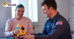 A friendly Fetch‑A‑Tech technician in uniform sits with a smiling customer at a cozy table in Las Vegas, offering her a cheerful dog-shaped plush toy. The Fetch‑A‑Tech plumbing and heating logo appears in the corner, highlighting the company’s caring, family-owned service dedicated to your comfort and peace of mind.