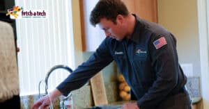 A friendly Fetch‑A‑Tech technician named Dennis, dressed in uniform, carefully inspects a kitchen sink faucet inside a comfortable Las Vegas home. The Fetch‑A‑Tech Plumbing, Heating & Air logo in the corner highlights our family-owned commitment to helpful service and your home's comfort.