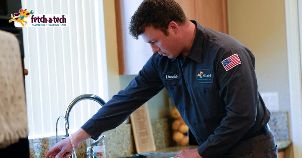A friendly Fetch‑A‑Tech technician named Dennis, dressed in uniform, carefully inspects a kitchen sink faucet inside a comfortable Las Vegas home. The Fetch‑A‑Tech Plumbing, Heating & Air logo in the corner highlights our family-owned commitment to helpful service and your home's comfort.
