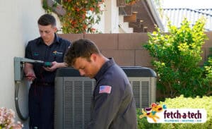 Two friendly Fetch‑A‑Tech technicians service an outdoor air conditioning unit beside a Las Vegas home, ensuring comfort and peace of mind for local families. The Fetch‑A‑Tech Plumbing, Heating & Air logo appears in the corner, highlighting our family-owned commitment to honest, helpful service.