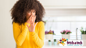 A woman in a yellow sweater covers her nose, reacting to an unpleasant smell in her home. The Fetch‑A‑Tech Plumbing, Heating & Air logo appears in the bottom right corner, highlighting our commitment to helping Las Vegas families enjoy clean air and greater comfort through honest, family-owned service.