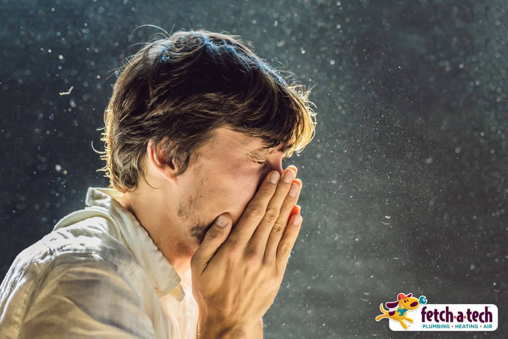 A man sneezes into his hands, with airborne particles visible, highlighting the importance of clean indoor air for your family’s comfort. The Fetch‑A‑Tech Plumbing, Heating & Air logo appears in the corner, representing our friendly, family-owned team proudly serving Las Vegas homes with honest and helpful service.
