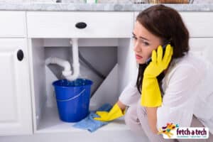 A woman in yellow cleaning gloves kneels beside a leaking kitchen sink pipe, calling for assistance as water drips into a blue bucket. She looks relieved to connect with Fetch‑A‑Tech’s friendly Las Vegas team—ready to provide honest, family-owned service and restore comfort to her home.