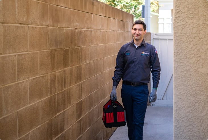 A friendly Fetch‑A‑Tech technician in uniform walks down a cozy Las Vegas outdoor corridor, carrying a tool bag and wearing work gloves—ready to help make this home more comfortable with expert insulation service.