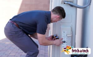 A friendly Fetch‑A‑Tech technician, dressed in a dark uniform, carefully inspects an outdoor unit to ensure your home stays comfortable. The Fetch‑A‑Tech Plumbing, Heating & Air logo highlights our reliable, family-owned service proudly serving the Las Vegas community.