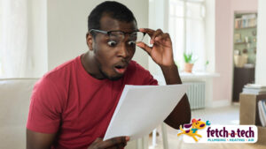 A friendly man in a red shirt reviews a document in his comfortable Las Vegas living room, looking pleasantly surprised by the helpful service information. The Fetch‑A‑Tech Plumbing, Heating, Air logo is displayed in the bottom right corner, reflecting our commitment to honest, family-owned service.