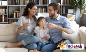 A smiling family—a man, woman, and young girl—relaxes together on their sofa in Las Vegas, enjoying refreshing drinks and the comfort of their home. The Fetch‑A‑Tech Plumbing, Heating & Air logo is featured in the corner, reflecting our commitment to honest, helpful service for local families.