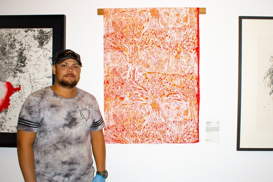 Jaivan is smiling next to an artwork made by his grandmother Madge Bowen. The artwork is an intricate red, orange and white screen print.