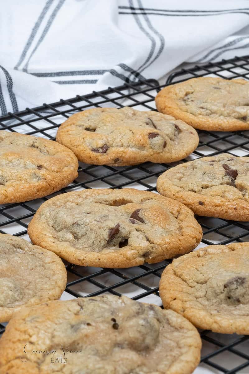 A cooling rack holding freshly baked chocolate chip cookies, made from the best chocolate chip cookie recipe without butter. The cookies appear golden brown with visible chocolate chunks, and a black and white striped cloth in the background adds a touch of elegance.