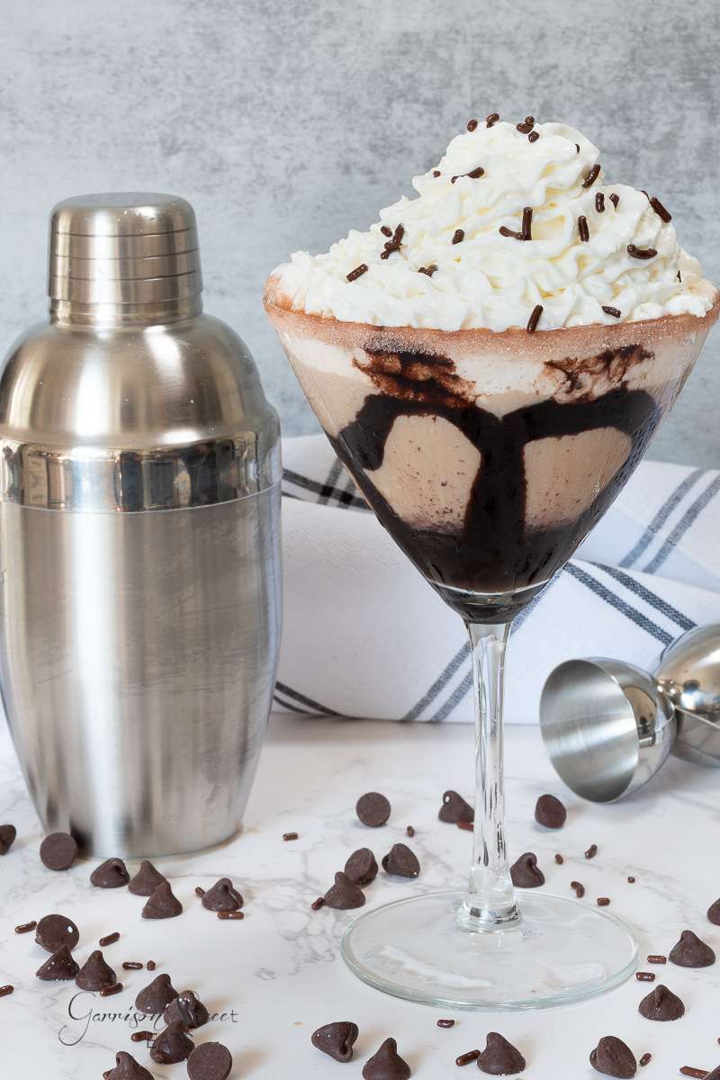 A chocolate martini in a cocktail glass with a chocolate swirl and topped with whipped cream and chocolate sprinkles is placed on a table. Surrounding the glass are chocolate chips, a metal cocktail shaker, and part of a striped cloth.