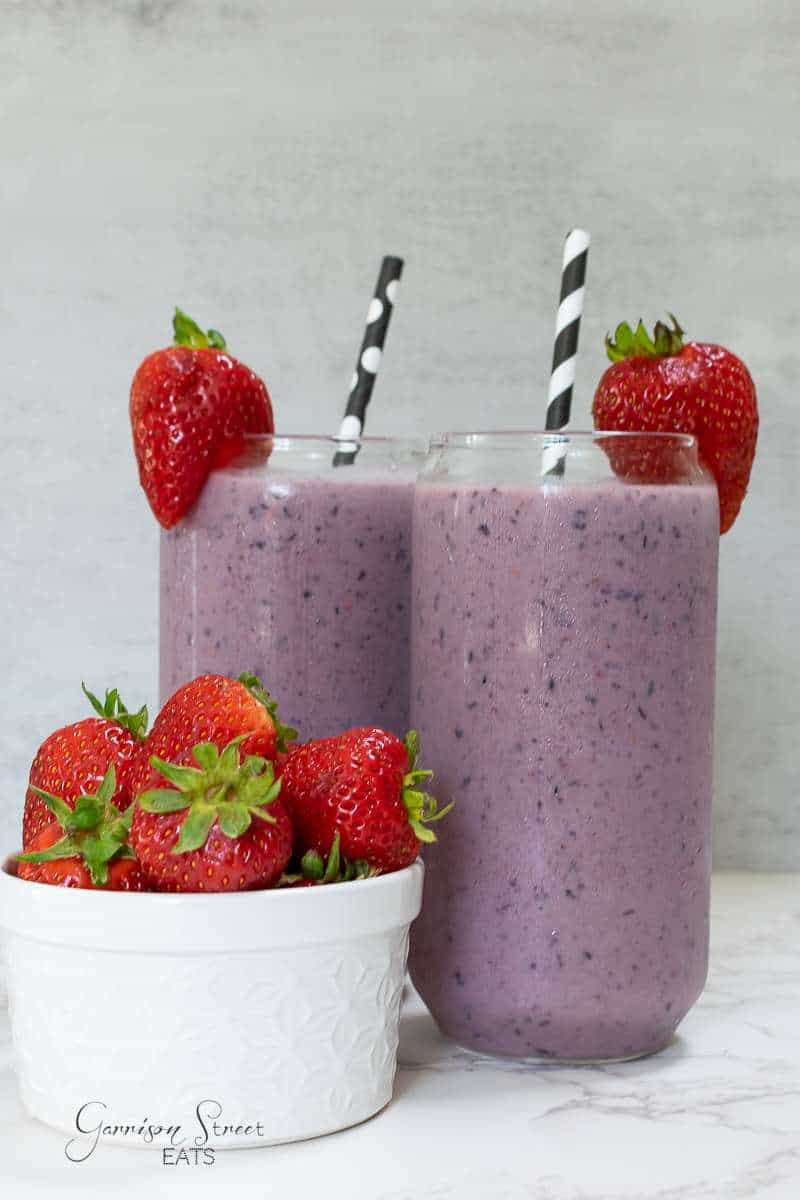 Two tall glasses of purple smoothie with black and white striped straws, each garnished with a fresh strawberry. A small white bowl filled with fresh strawberries sits next to the glasses on a light textured surface.