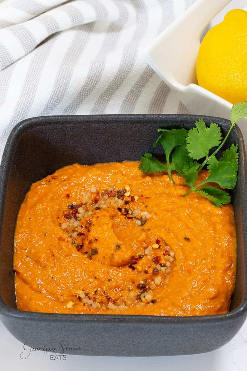 A square black bowl containing a vibrant orange-colored hummus, garnished with red pepper flakes and a sprig of cilantro. In the background, there is a yellow lemon in a white bowl, all placed on a white and gray-striped cloth.