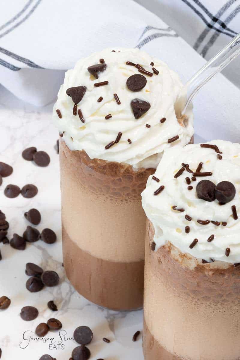 Two tall glasses are filled with a frothy Chocolate Chip Mocha Frappuccino, topped with whipped cream, chocolate chips, and chocolate sprinkles. A clear straw extends from one of the glasses. Chocolate chips are scattered around on the white marble surface.