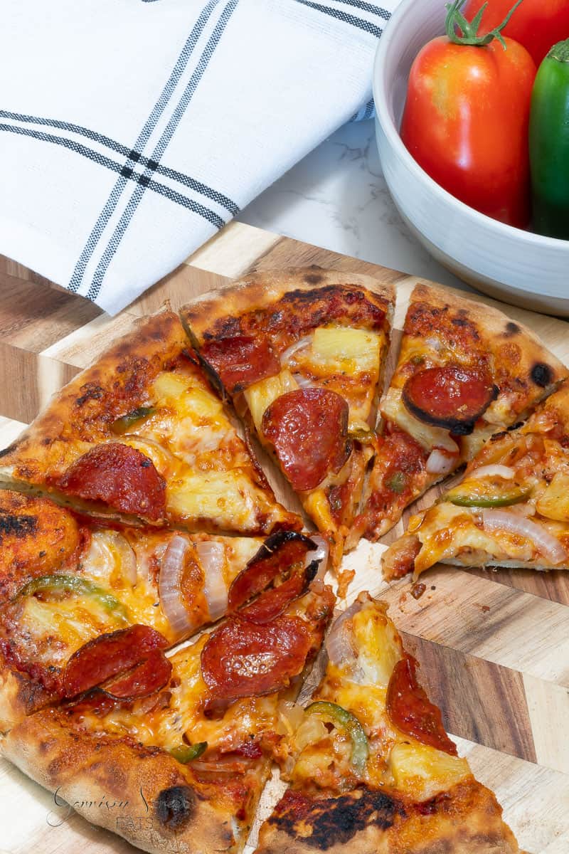 A sliced pizza on a wooden board, topped with pepperoni, pineapple, onions, and jalapeno peppers showcases how to make Hawaiian Pepperoni and Pineapple Pizza. In the background, a white dish contains tomatoes and a jalapeno. A white and blue striped cloth towel also sits nearby.