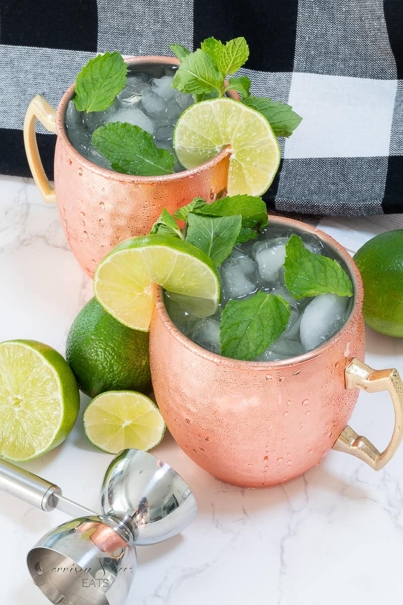 Two copper mugs filled with a cocktail on the rocks, garnished with fresh mint leaves and lime slices. A few whole limes and lime slices are scattered around, along with a metal cocktail jigger. A black and white checkered cloth is in the background, complimenting the best ginger beer for Moscow Mules.